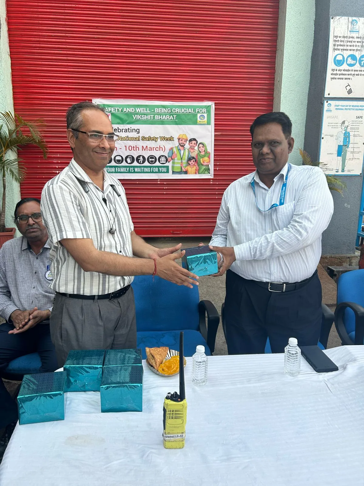 Two participants shake hands over a presentation item in front of a red roller shutter and a safety poster.