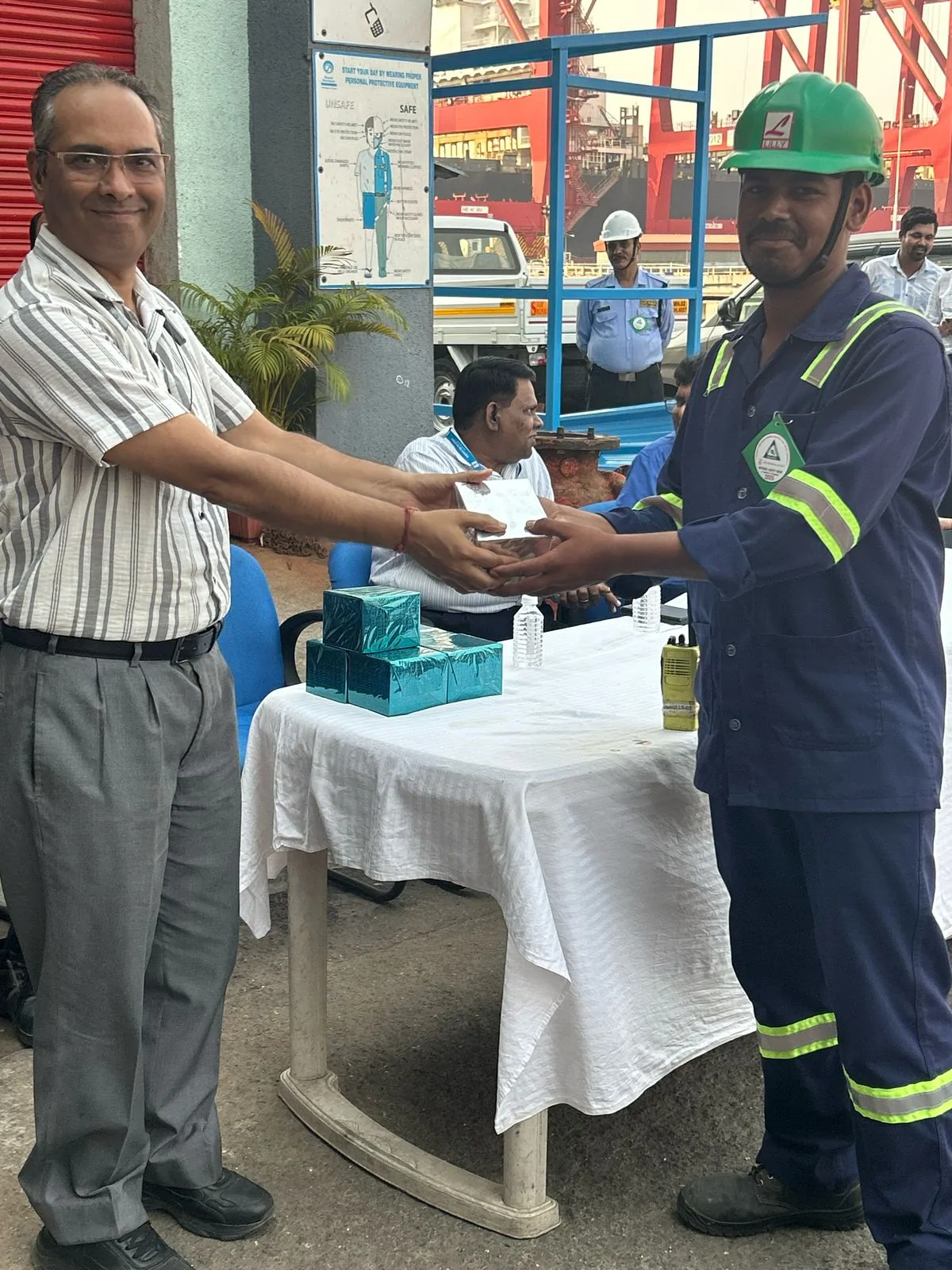 A worker in navy coveralls, reflective strips and a green hard hat receives a presentation item at a terminal gathering.