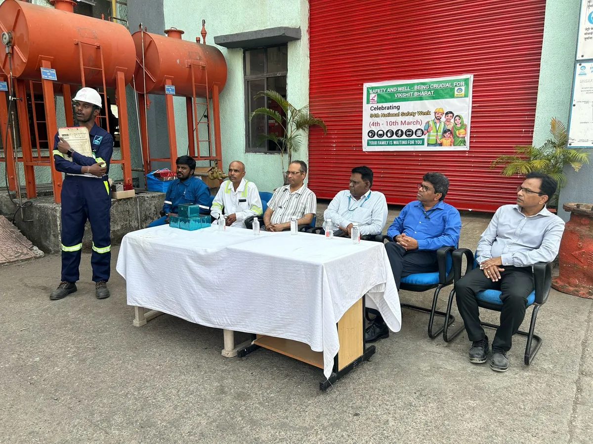 A speaker wearing a hard hat addresses seated participants beside red fire equipment and a white-covered table.