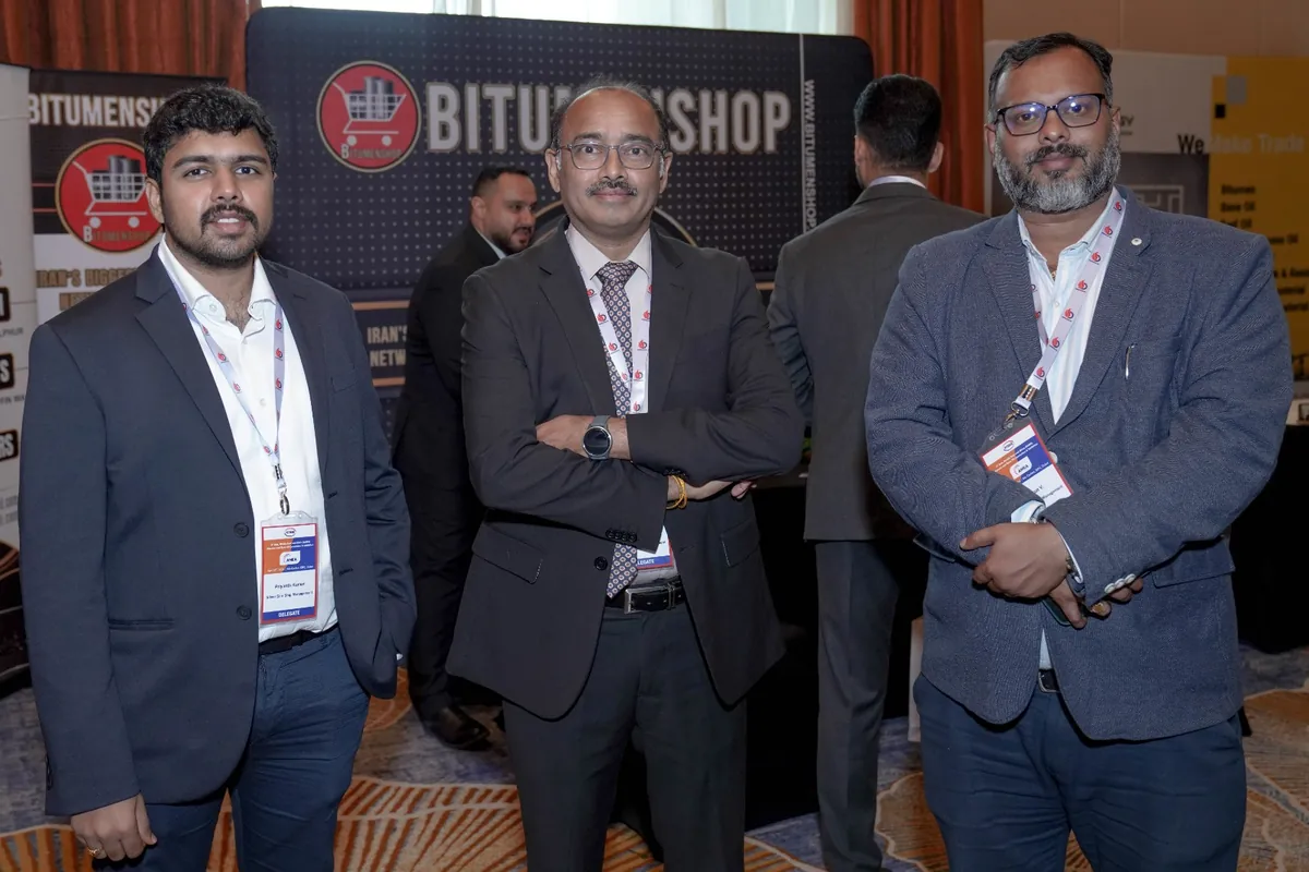 Three suited delegates wearing lanyards pose together in front of a Bitumenshop exhibition stand.