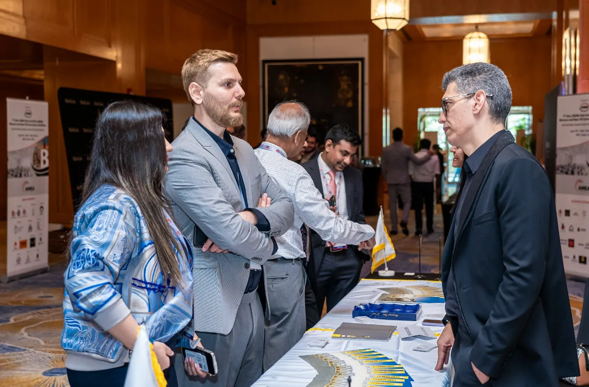 Three attendees talk across an exhibition table displaying brochures at an industry event.