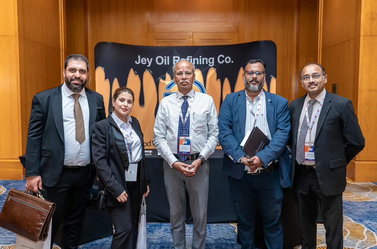 Five delegates wearing conference lanyards pose in front of a Jey Oil Refining Co. display.