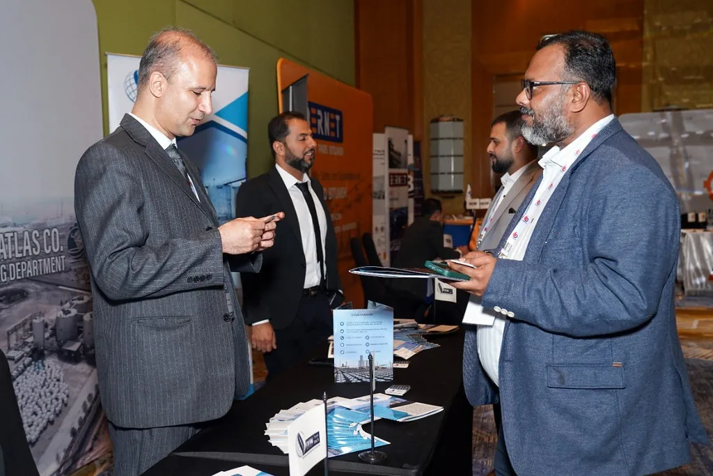 Two suited attendees exchange information beside brochures at an exhibition stand.