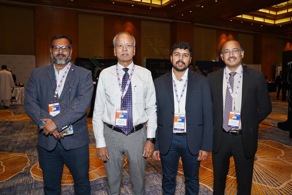 Four delegates wearing conference badges pose together in a hotel event hall.