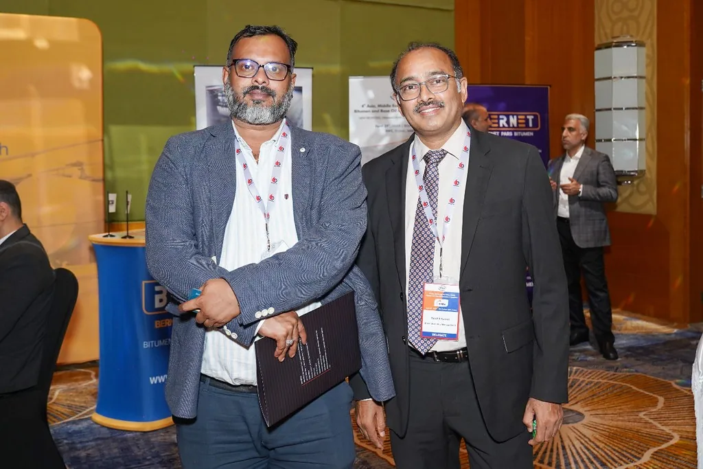 Two suited delegates wearing event lanyards pose together beside a conference exhibition area.