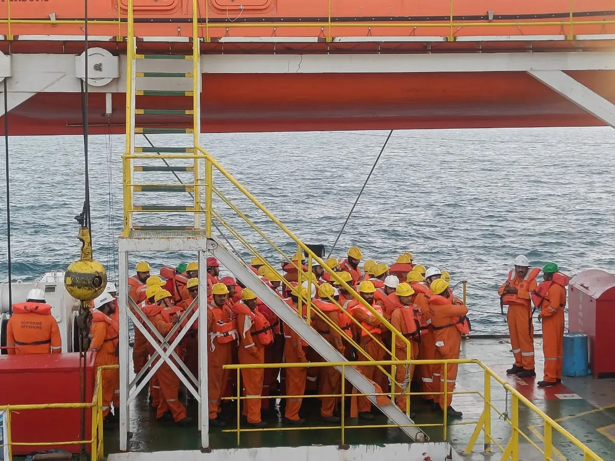 Crew wearing orange lifejackets and hard hats assemble on a vessel deck beneath an orange enclosed lifeboat.