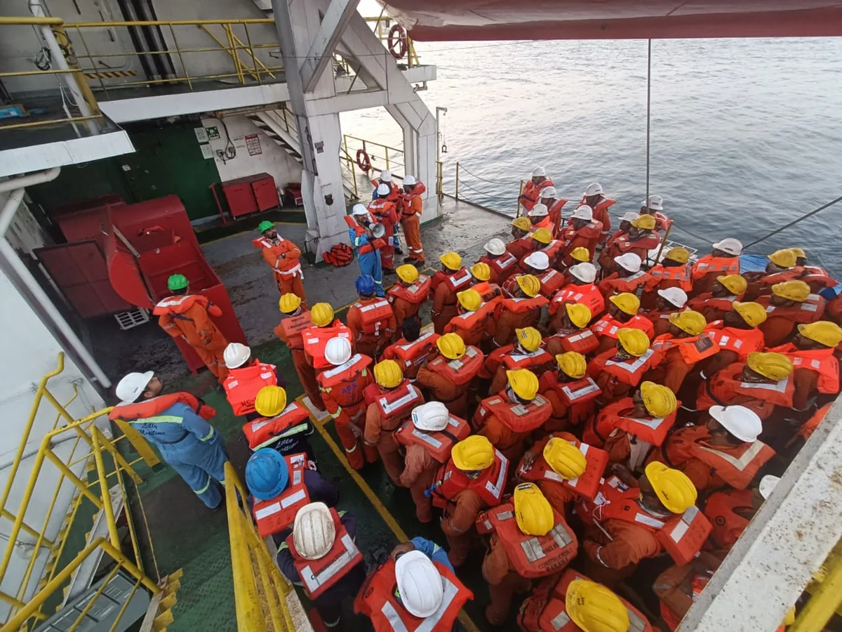 An overhead view shows a large crew in orange lifejackets and yellow or white hard hats gathered beside a vessel railing.