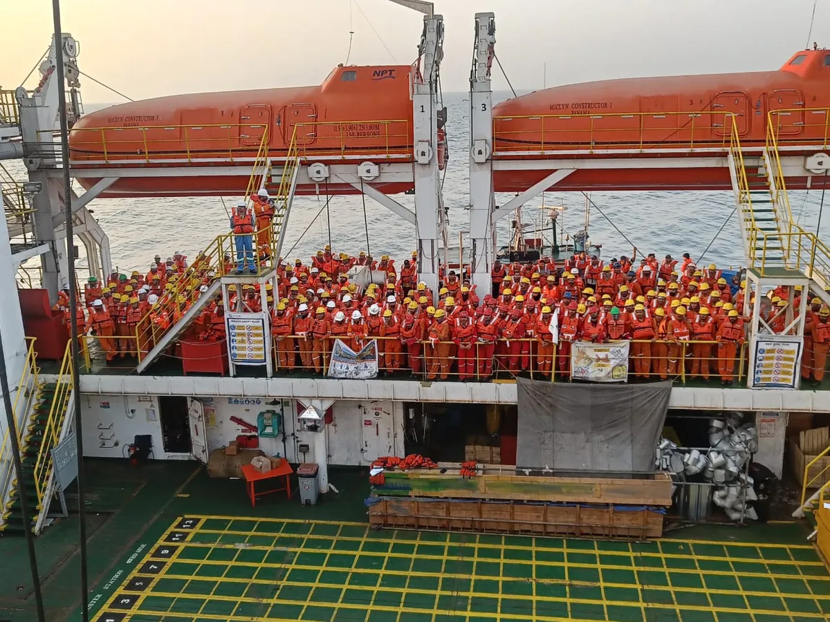 A large group of crew in orange lifejackets stands on the deck beneath two orange enclosed lifeboats.