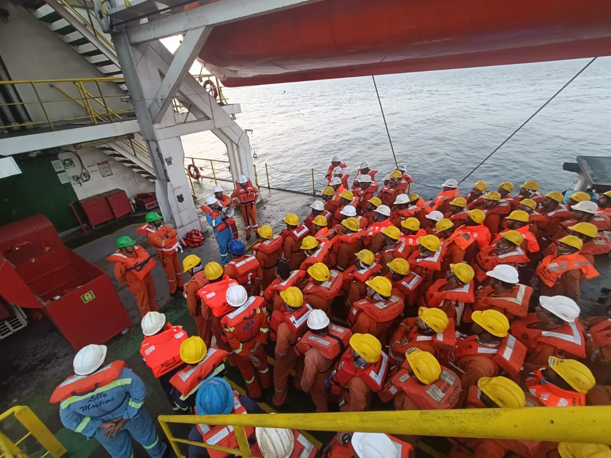 Crew in orange lifejackets and hard hats gather between a vessel superstructure and the sea-facing railing.