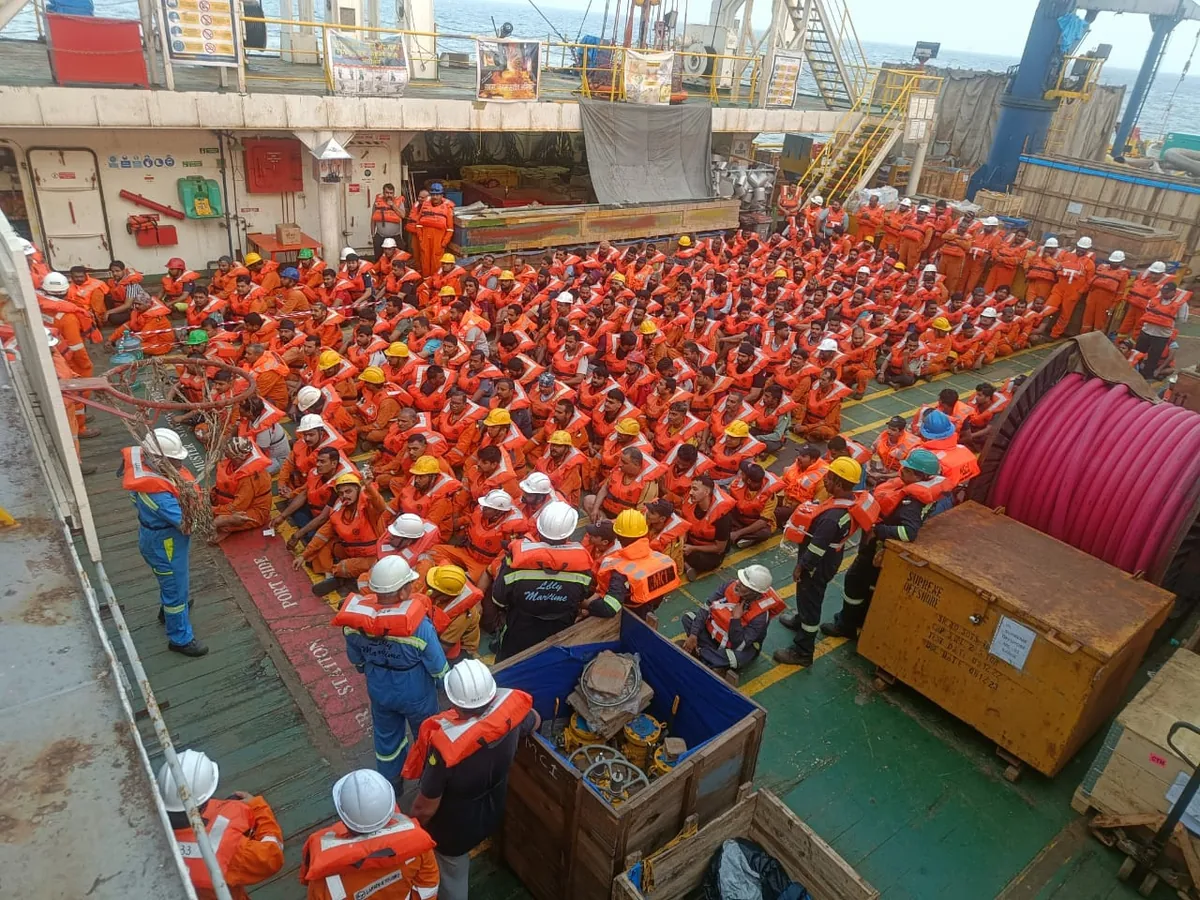 A high-angle view shows a crowded vessel working deck filled with crew in orange lifejackets and hard hats.