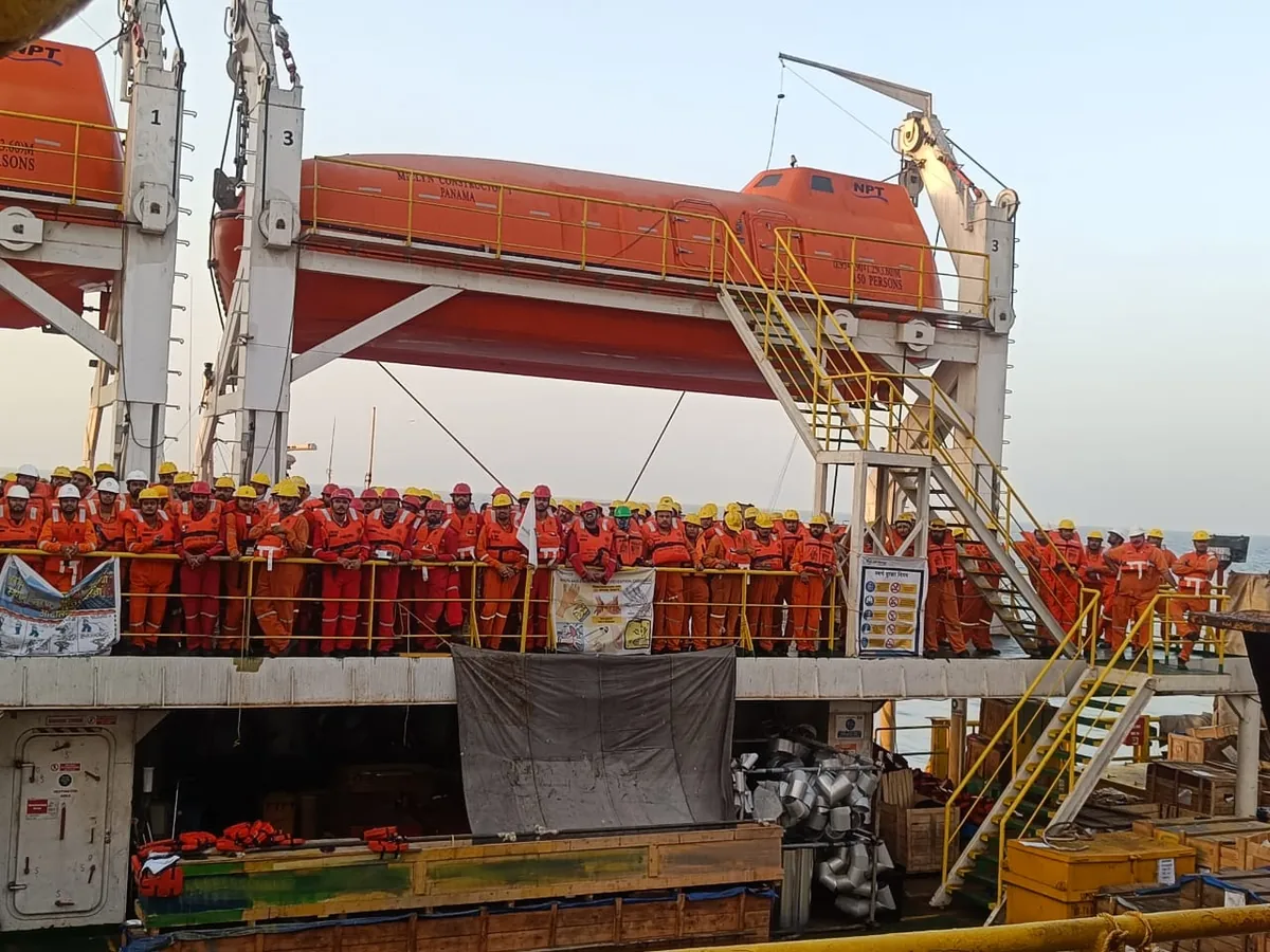A side view shows crew assembled below an orange enclosed lifeboat and a yellow access stairway.