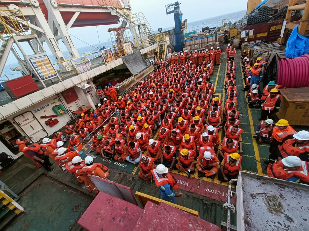 Crew in orange lifejackets stand in rows along a vessel deck beside coiled lines, deck equipment and the accommodation block.