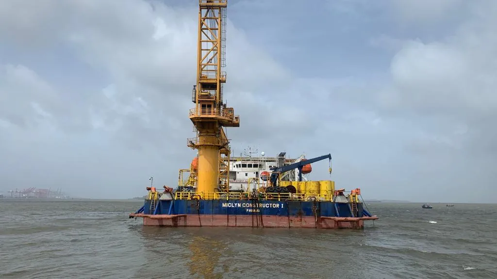 A large yellow crane rises above the bow end of an operational vessel on grey water beneath heavy clouds.