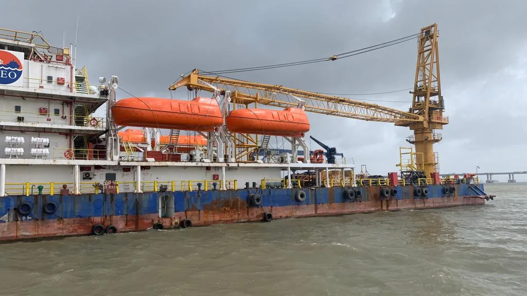 The side of a crane vessel shows orange enclosed lifeboats, white accommodation decks and a yellow crane above a blue hull.