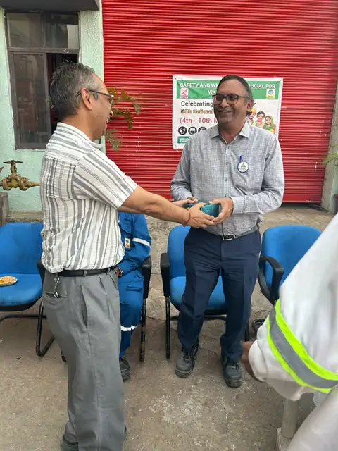 Two men exchange a presentation item beside blue chairs and a red roller shutter at a terminal safety gathering.