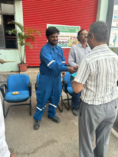 A worker in blue coveralls receives a presentation item beside seated participants and a red roller shutter.