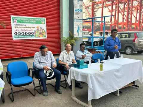 A man in blue workwear speaks beside a white-covered table while participants sit in front of a red roller shutter.