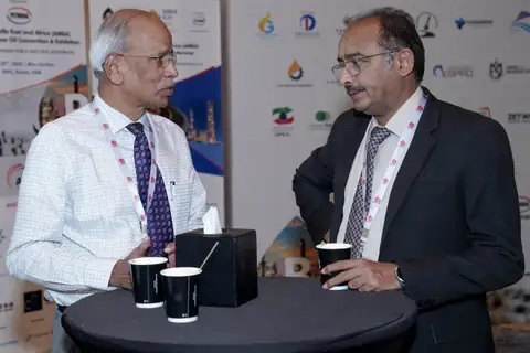 Two delegates with event lanyards talk beside a round table holding coffee cups and presentation boxes.