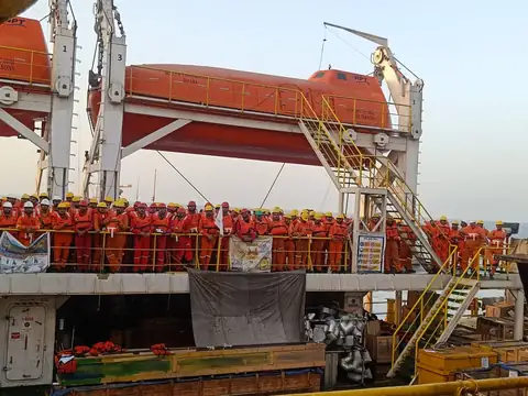 A side view shows crew assembled below an orange enclosed lifeboat and a yellow access stairway.