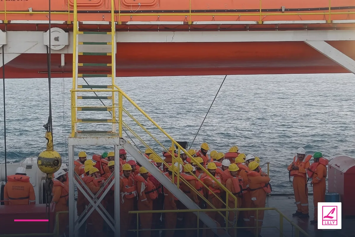 Crew in hard hats stand on deck beneath an orange enclosed lifeboat, with a navy and magenta Lilly Maritime treatment.